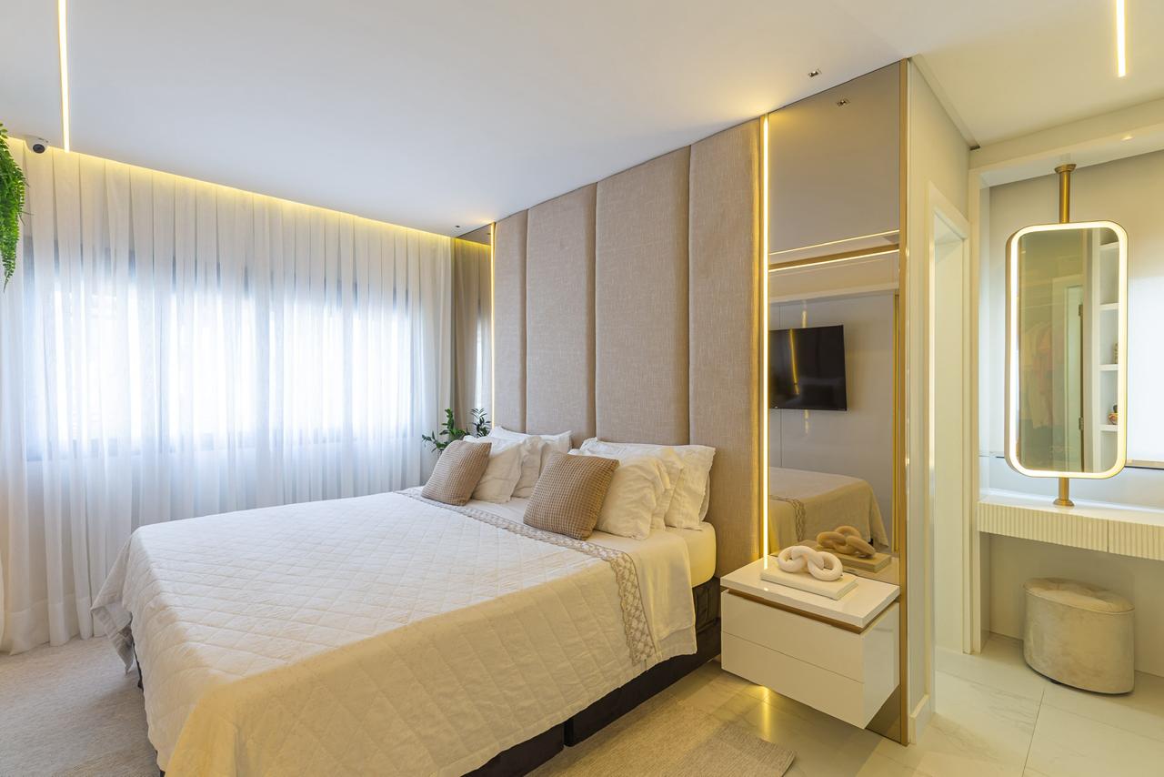 Suíte em tons neutros com cama clara, cortinas leves e iluminação quente